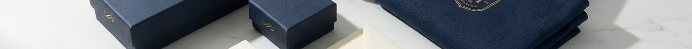 A flat lay of cohesive branded print materials including custom packaging boxes, business cards, a sealed envelope, and a tote bag in a unified navy and gold color scheme on a marble surface.